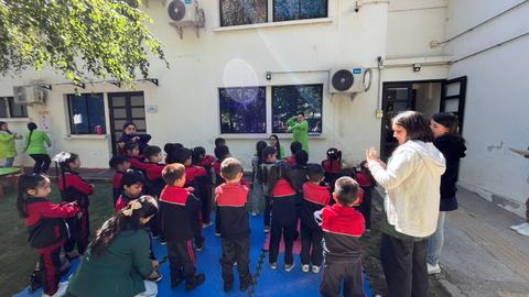 Un grupo de niños en un patio escolar escucha a un adulto que les está enseñando algo.
