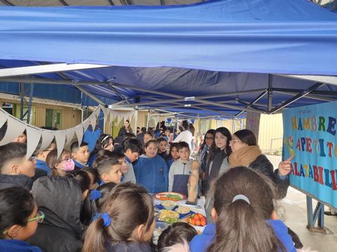Un grupo de estudiantes observa una actividad con alimentos en un evento escolar bajo una carpa.