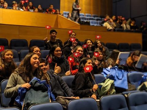 Un grupo de jóvenes sonrientes sostienen banderas en un evento en un auditorio.