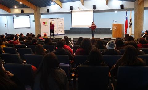 Una presentación en un auditorio con un grupo de personas sentadas frente a una pizarra y oradoras en el escenario.