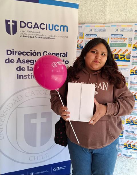 Una persona sostiene un dispositivo en una mano y un globo rosa en la otra, frente a un fondo con un cartel de una universidad.