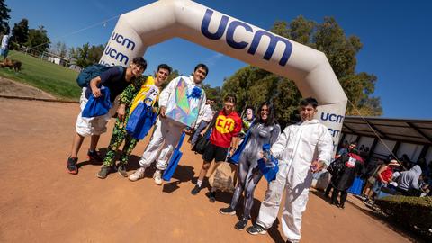 Un grupo de jóvenes vistiendo disfraces coloridos se reúne en un evento al aire libre bajo un arco con el logo de UCM.