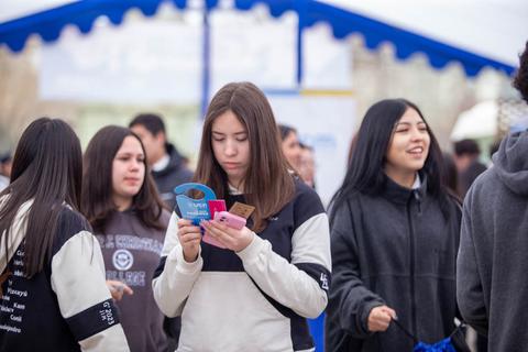 Un grupo de jóvenes en un evento al aire libre, donde una chica está revisando unos papeles en su mano.