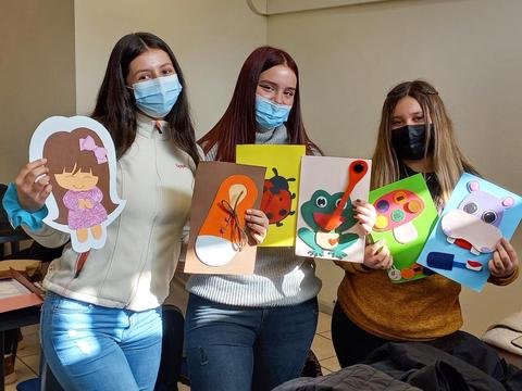 Tres jóvenes sostienen manualidades coloridas en un ambiente de aula.