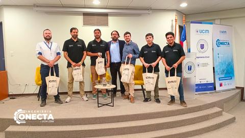 Un grupo de jóvenes con camisetas negras posa en un evento en una sala con banderas y un banner de UCM.