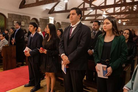 Un grupo de personas vestidas de forma formal asisten a una ceremonia en un lugar con un ambiente solemne.