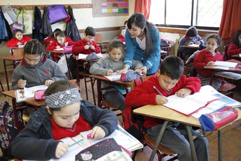Un aula escolar con niños en actividades de escritura y una maestra asistiendo.
