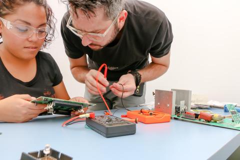 Dos personas están trabajando en la reparación de un circuito electrónico en una mesa de trabajo.