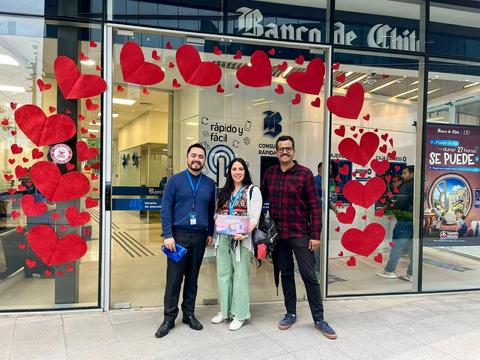 Un grupo de tres personas sonrientes posa frente a un banco decorado con corazones rojos.