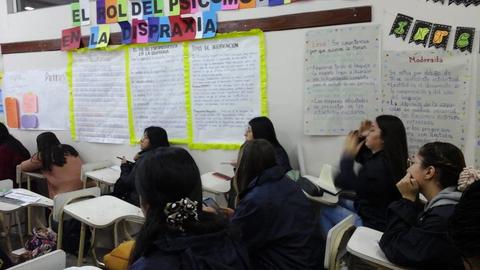 Una clase en un salón de estudios donde los estudiantes están sentados en pupitres, rodeados de carteles informativos en la pared.