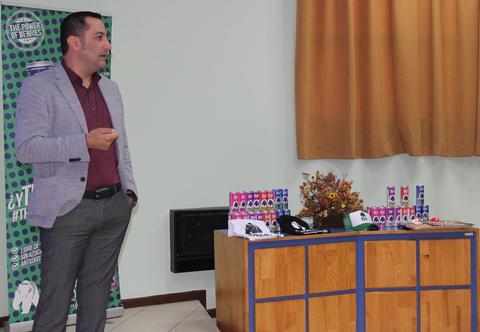 Un hombre de pie, hablando frente a una mesa con diversos productos y decoraciones en un salón.