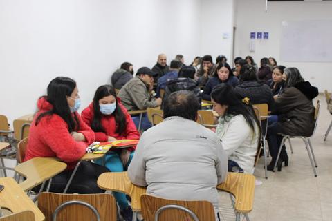 Un grupo de personas se reúne en un aula, conversando en pequeños grupos alrededor de mesas.