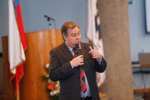 Un hombre de pie en un escenario, hablando con un micrófono en la mano y un fondo con banderas.