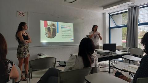 Una presentación en una sala con un hombre hablando frente a una pantalla y una mujer observando.