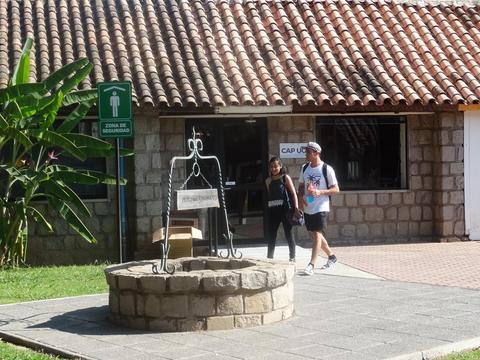 Dos personas caminan cerca de un pozo de agua en un entorno al aire libre con un edificio de fondo.