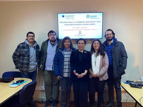 Un grupo de personas se encuentra en una sala de reuniones junto a una pantalla que presenta información sobre visas migratorias y el fenómeno 'brain waste' en la migración hacia Estados Unidos.