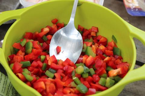 Un recipiente grande de color verde contiene una mezcla de pimientos rojos y verdes picados, con una cuchara de metal en su interior.