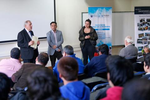 Un grupo de personas asiste a una conferencia en una sala con un presentador y dos panelistas en el escenario.
