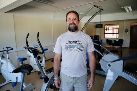 Un hombre sonriente está de pie en un gimnasio, rodeado de bicicletas estáticas y otros equipos de ejercicio.