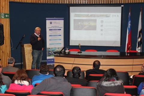 Un conferencista está hablando frente a una audiencia en un aula con una pantalla detrás.