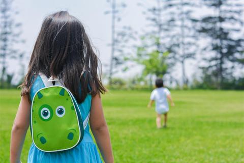 Una niña con una mochila de dinosaurio observa a un niño que corre en un campo verde.