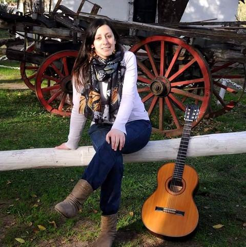 Una mujer sentada sobre un tronco junto a una guitarra en un paisaje rural.