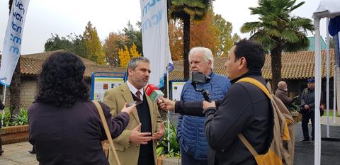 Un grupo de personas realiza una entrevista en un evento al aire libre rodeado de plantas y árboles.