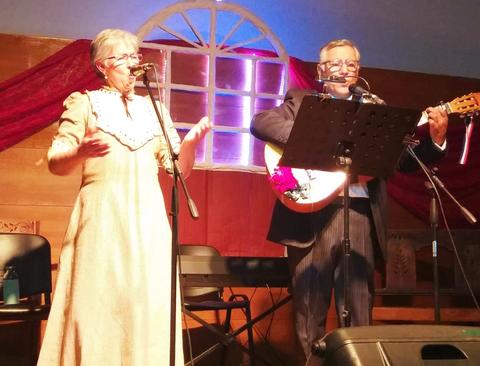 Una pareja de músicos actuando en un escenario con micrófonos y un fondo decorativo.