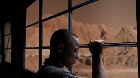 Un hombre mira por una ventana hacia un paisaje árido y montañoso de color marrón.