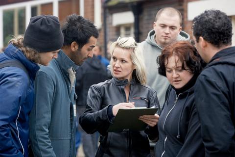 Un grupo de seis personas conversa intensamente en una calle mientras una mujer toma notas en una carpeta.