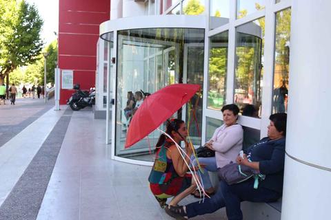 Tres mujeres conversan en la entrada de un edificio, una de ellas sostiene un paraguas rojo.