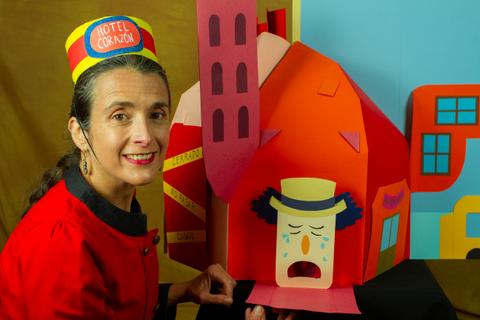 Una mujer sonriente vestida de manera colorida al lado de una casa de papel con una expresión triste.