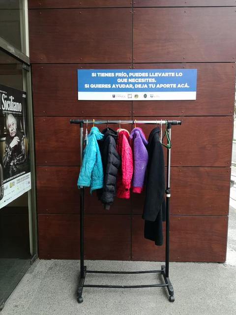 Un perchero con abrigos de distintos colores está expuesto en la calle, junto a un mensaje que invita a las personas a llevarse lo que necesiten y a donar si desean ayudar.