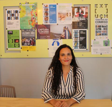 Una mujer sonriente con camisa de rayas se encuentra sentada frente a una pared llena de carteles y promociones.