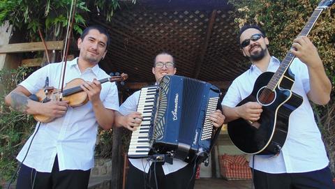 Tres músicos sonrientes posan con sus instrumentos en un entorno al aire libre.