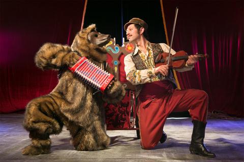 Un hombre vestido con ropa colorida toca el violín mientras un oso de peluche toca el acordeón en un espectáculo circense.