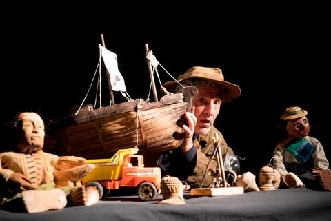 Un hombre presenta una serie de figuras y un barco de madera sobre una mesa oscura.