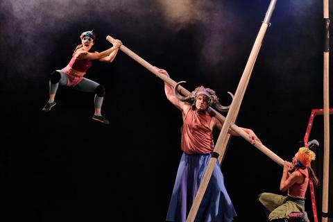 Una actuación acrobática con tres artistas que utilizan barras largas en un ambiente dramático y colorido.