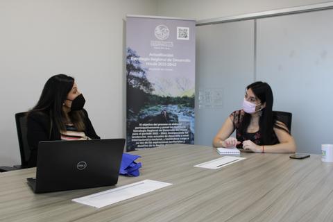 Dos mujeres conversan en una sala de reuniones con mascarillas mientras están sentadas frente a una mesa.