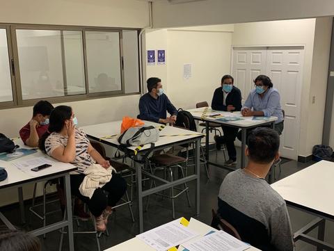 Un grupo de personas se encuentra en un salón de clases, algunas conversando mientras otras toman apuntes.