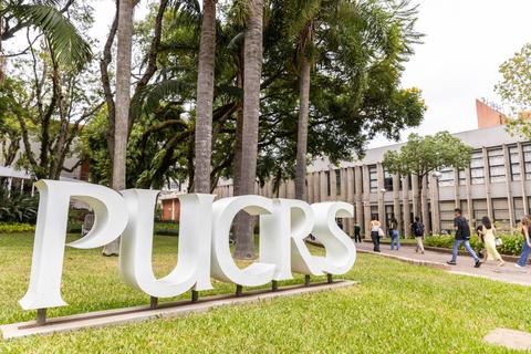 Imagen del campus de la PUCRS con un gran letrero que lleva sus iniciales y personas caminando a lo lejos.