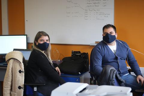 Dos personas sentadas en una oficina, usando mascarillas y frente a computadoras.
