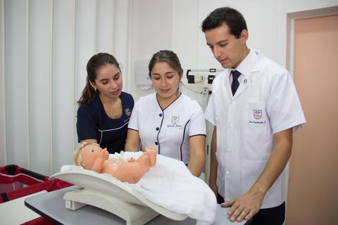 Tres profesionales de la salud observan a un bebé de práctica sobre una tabla en un entorno médico.