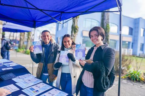 Tres personas sonrientes sostienen folletos bajo una carpa en un evento al aire libre.