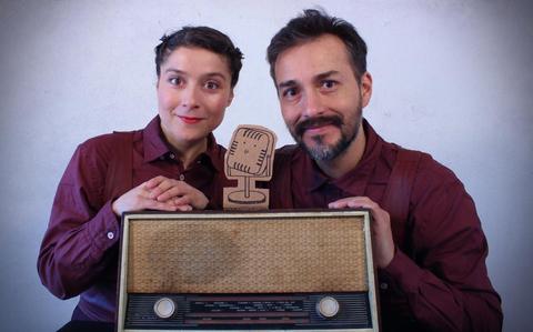 Una pareja sonriente sostiene un viejo radio con un micrófono de cartón delante de ellos.