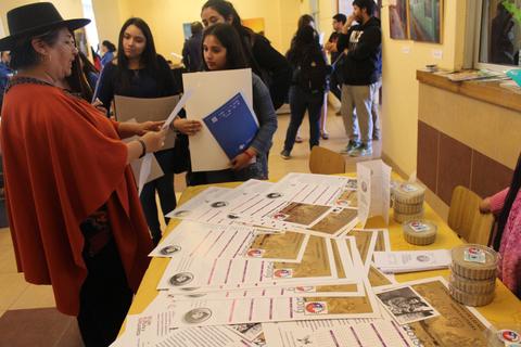 Una mujer con sombrero interactúa con un grupo en un evento educativo rodeado de documentos en una mesa.