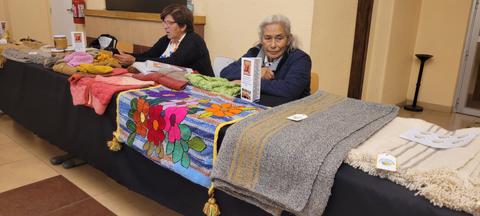 Dos mujeres están sentadas detrás de una mesa exhibiendo mantas de diversos colores y diseños.