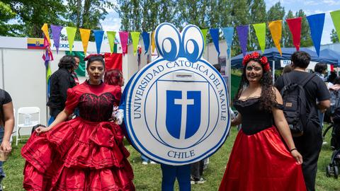 Dos mujeres vestidas con trajes tradicionales posan junto a un escudo en un evento al aire libre.