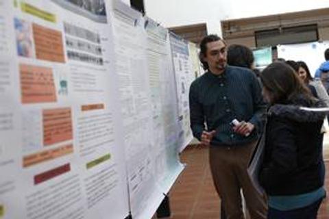 Un hombre está presentando un póster en un evento académico mientras una mujer lo escucha atentamente.