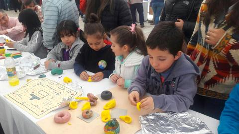 Un grupo de niños juega con plastilina en una mesa mientras otros participan en actividades detrás de ellos.